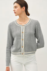 A woman models a grey cable knit cardigan with white trim and brown buttons.