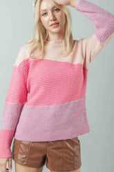 A blonde model wearing a crew-neck, color-blocked sweater with mixed knit textures, including a smooth pale nude shoulder and a ribbed vibrant pink chest. The look is completed with brown leather-look shorts