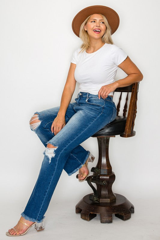 Woman smiling while wearing bytos distressed high rise bootcut jeans and a white top, seated on a wooden stool.