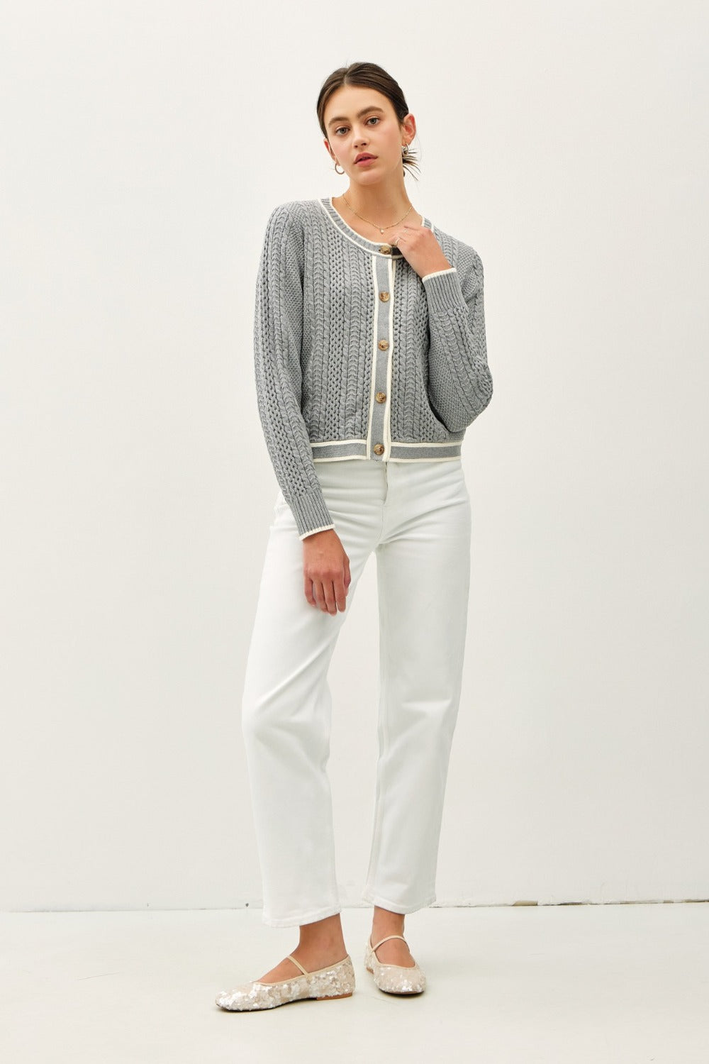 A front-facing, full-body view of a model wearing the grey cable knit cardigan to showcase its fit and length. She looks at the camera with one hand near her neckline.