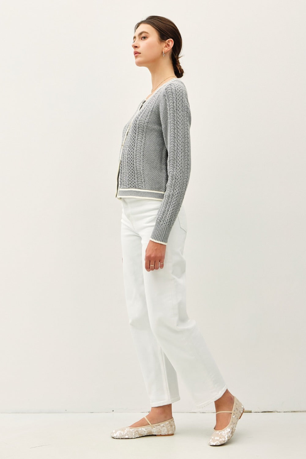 A model turned slightly to the side wears a grey cable knit long-sleeve cardigan with a contrast cream edge along the round neckline and button placket, paired with white pants.