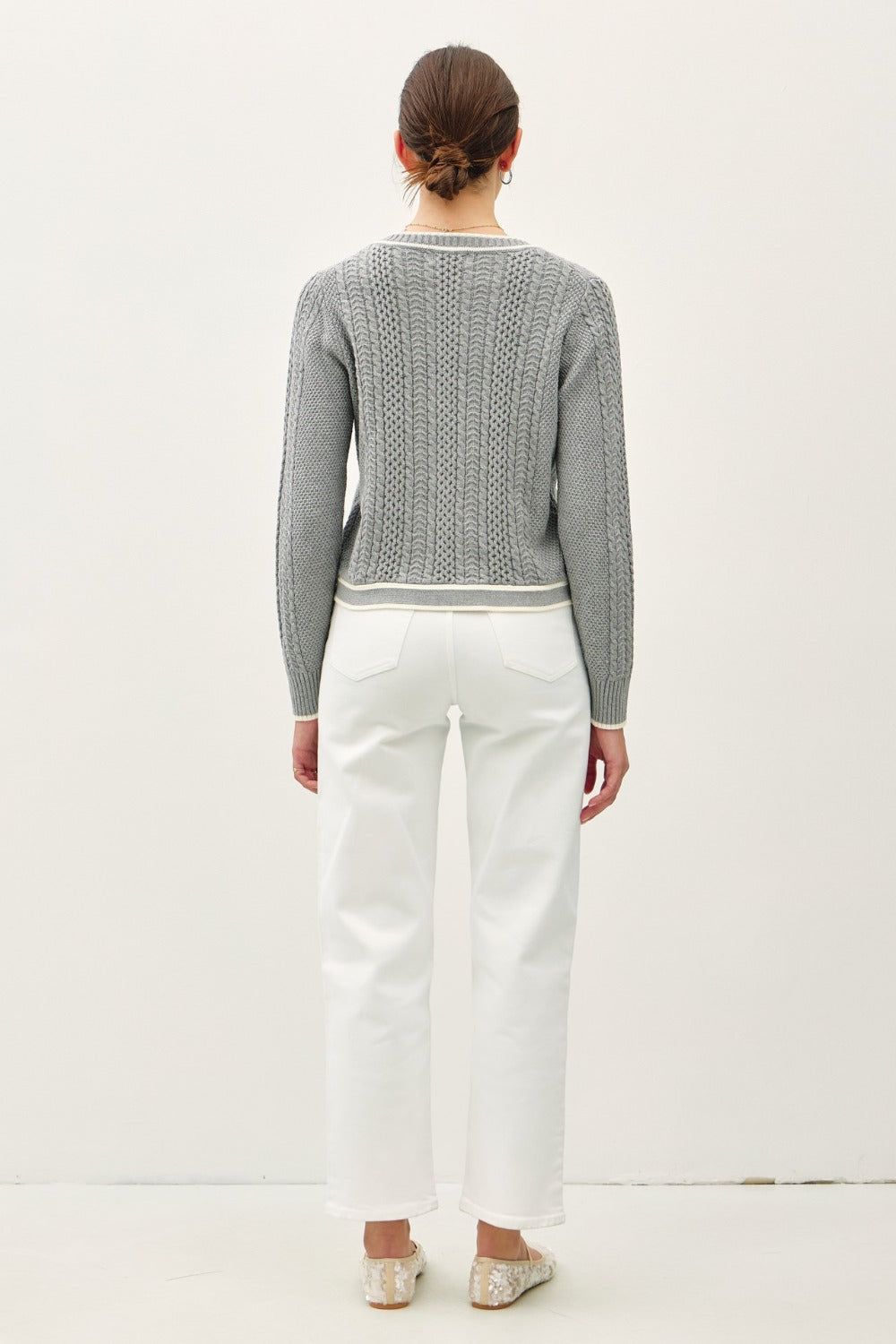 A woman with her hair tied back models a grey, long-sleeved cable knit cardigan. The sweater features a round neckline, brown buttons, and a contrasting off-white trim along the placket, cuffs, and hem.