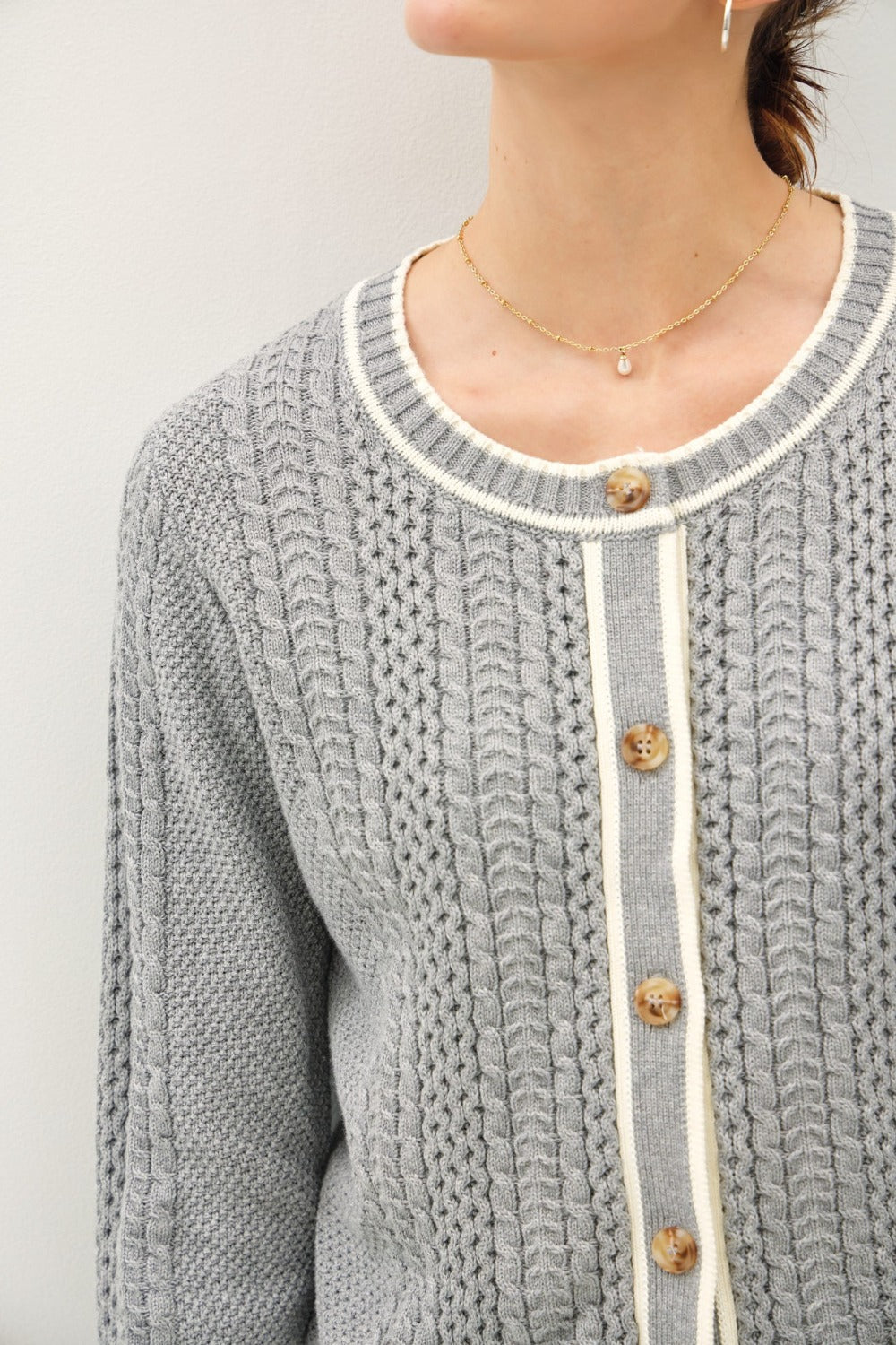 A detailed view of a grey cable knit cardigan, showing the intricate knit pattern, the round neckline with cream trim, and the light brown buttons on the placket.