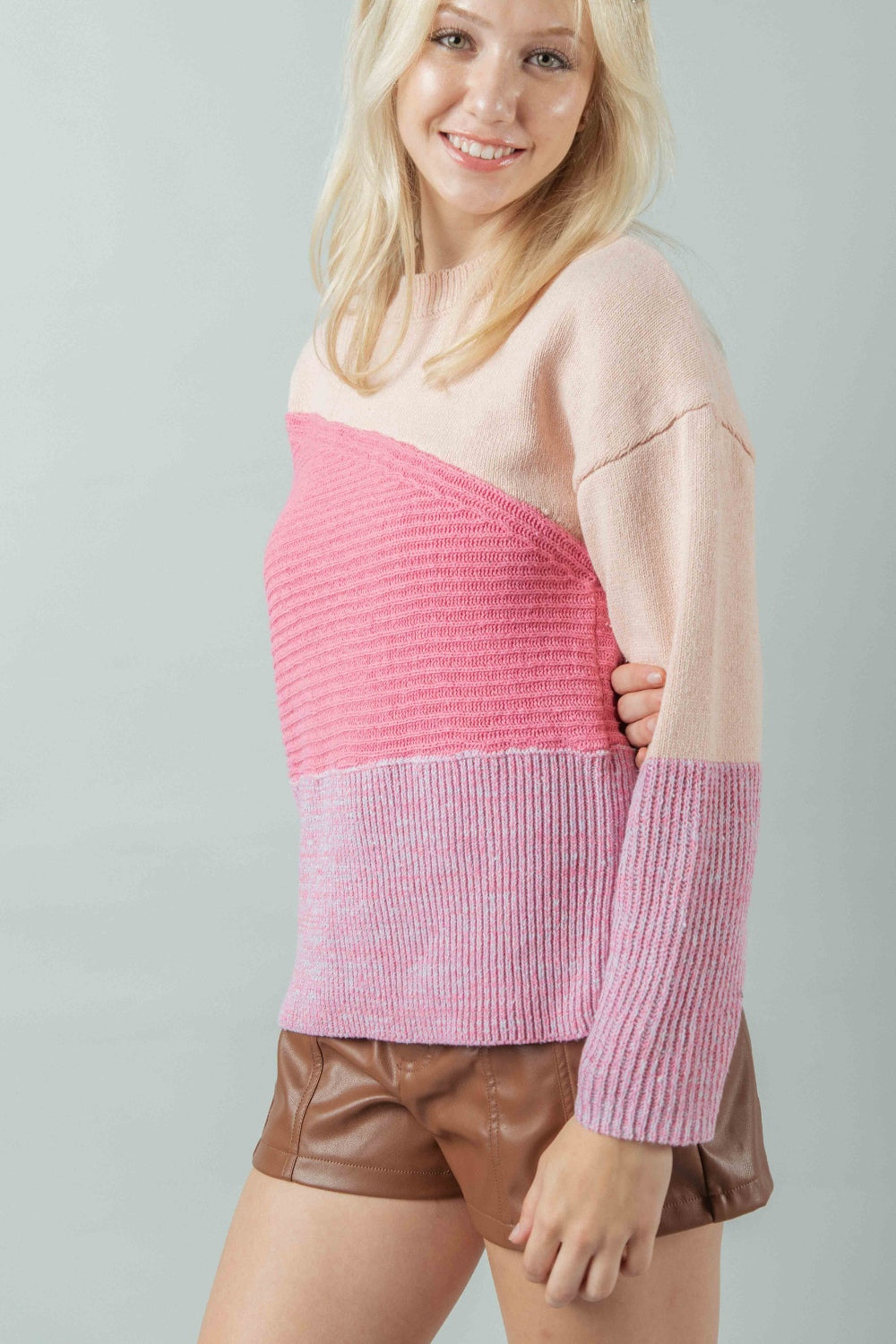 A Side shot of the model who is smiling wearing the color-blocked sweater to illustrate its profile and fit over brown leather-look shorts. The image clearly shows the contrasting knit textures.