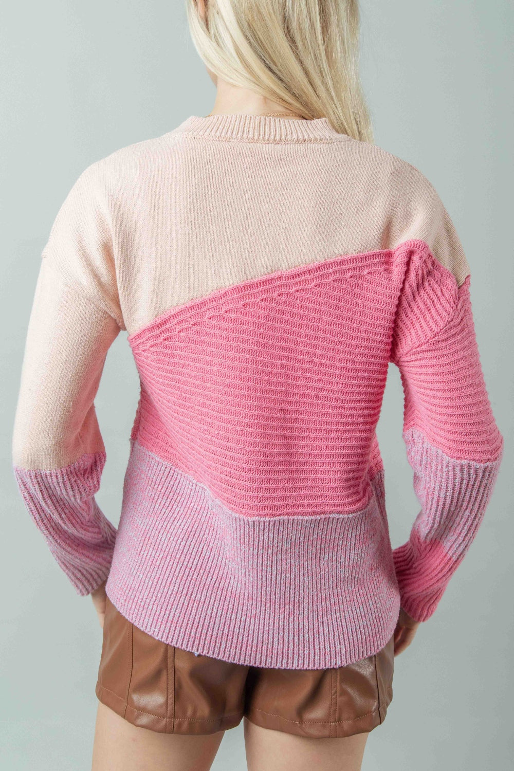 The back of a long-sleeved sweater worn with brown shorts, featuring a unique diagonal color block design with sections of smooth pale peach, ribbed hot pink, and textured heathered pink.